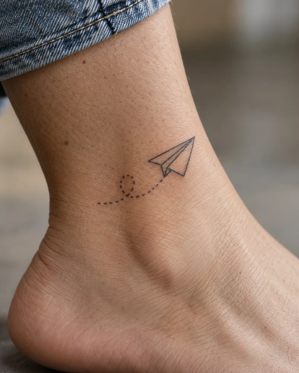 Fine-line paper plane tattoo and dotted trail on an ankle