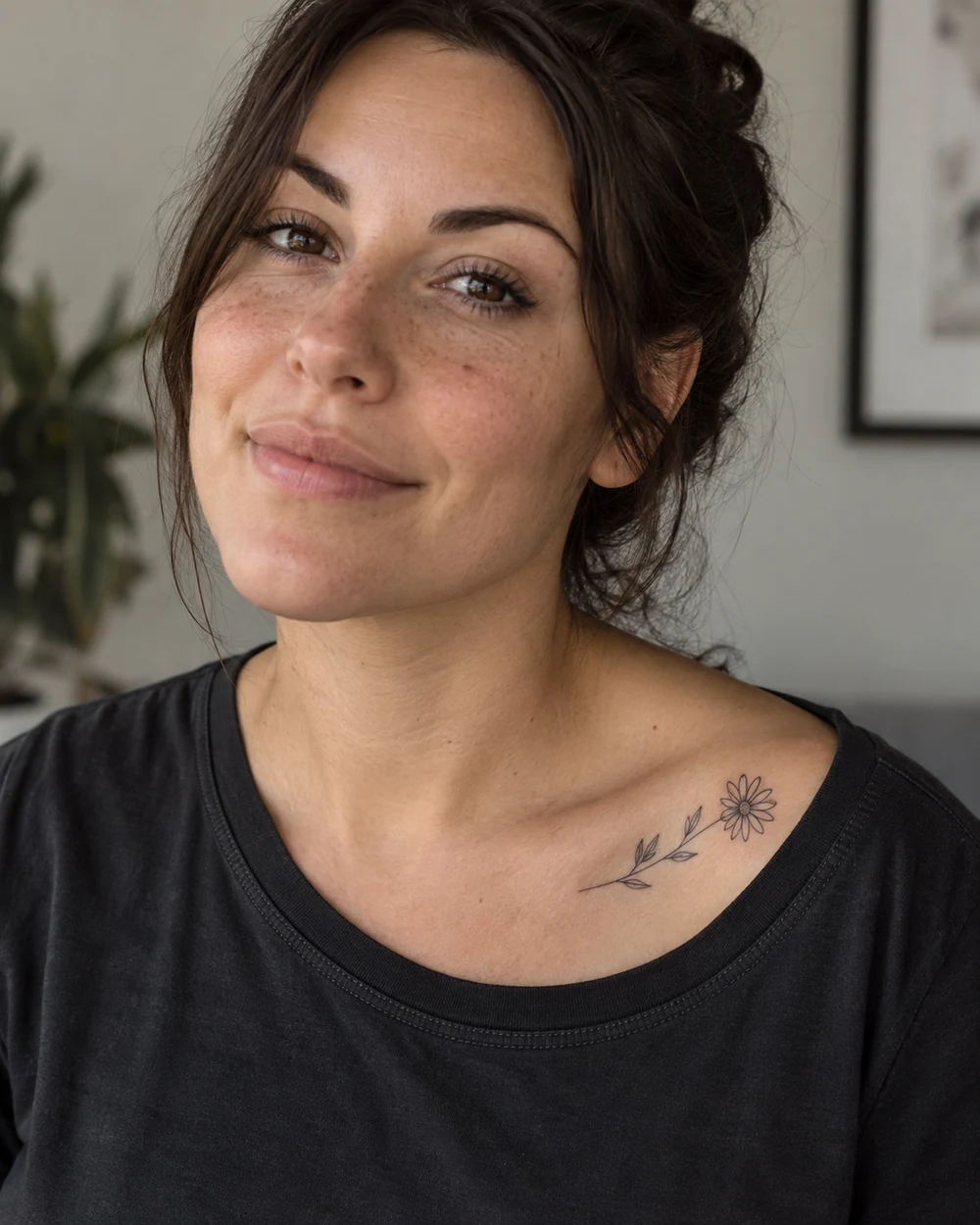 Small black fine-line daisy tattoo with a leafy stem along a woman's collarbone