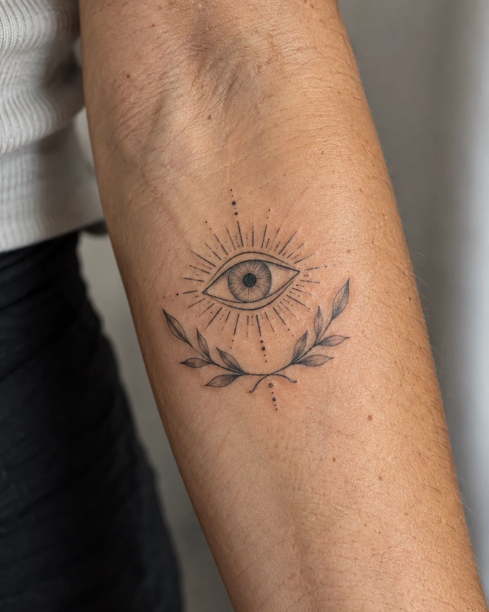 Fine-line evil eye tattoo above a curved leafy vine on a forearm