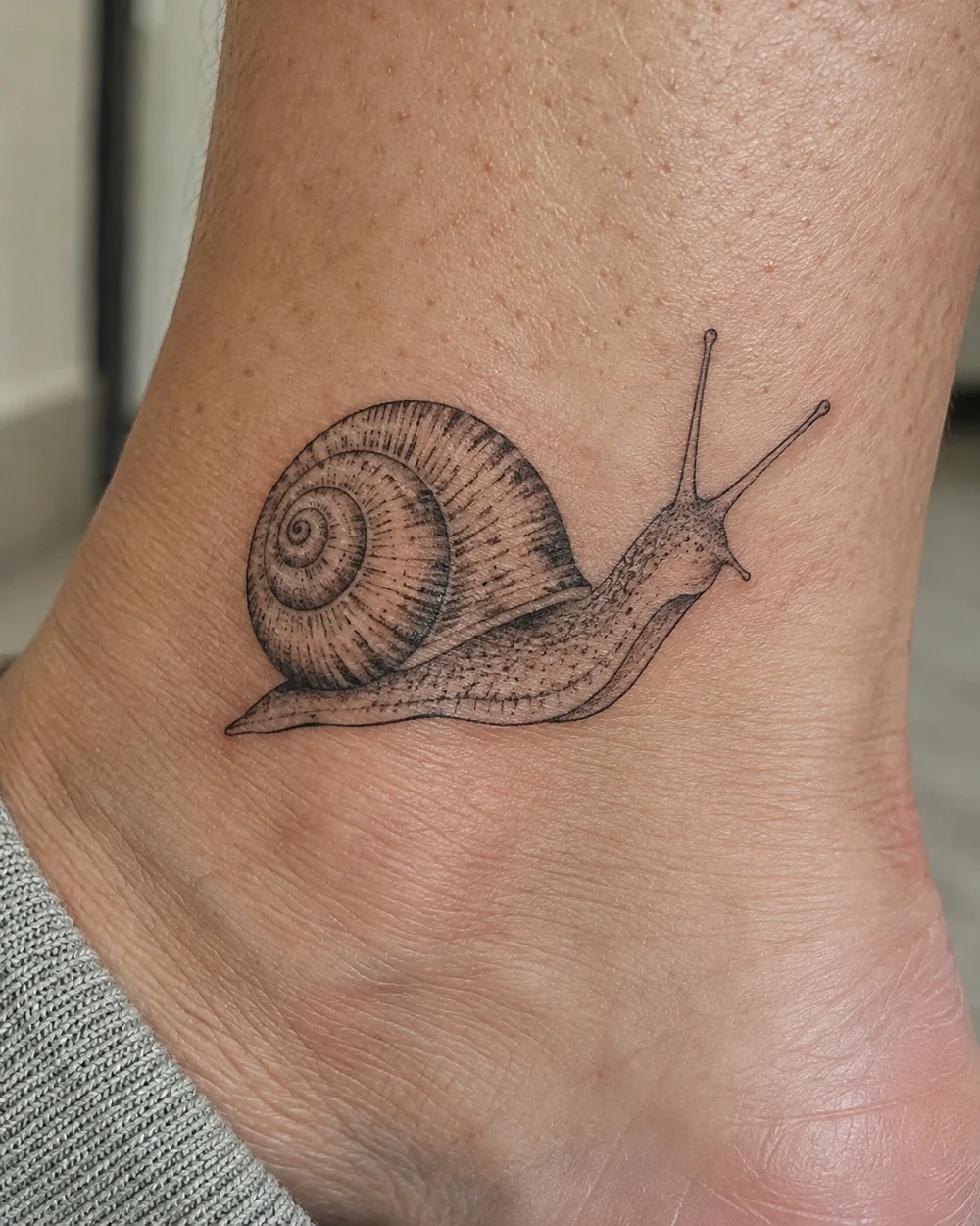 Fine line snail tattoo with spiral shell and two feelers on an ankle