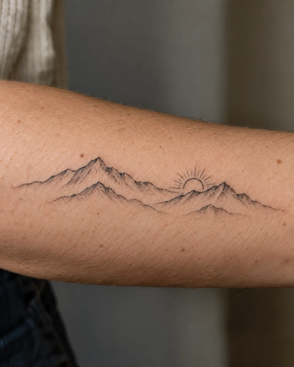 Two fine-line mountain ridges and a rising sun in a horizontal forearm tattoo