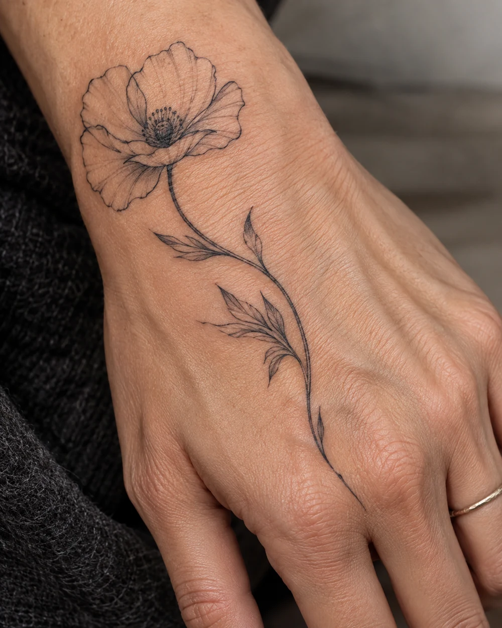 Fine-line open poppy tattoo with curved leafy stem across the back of a hand