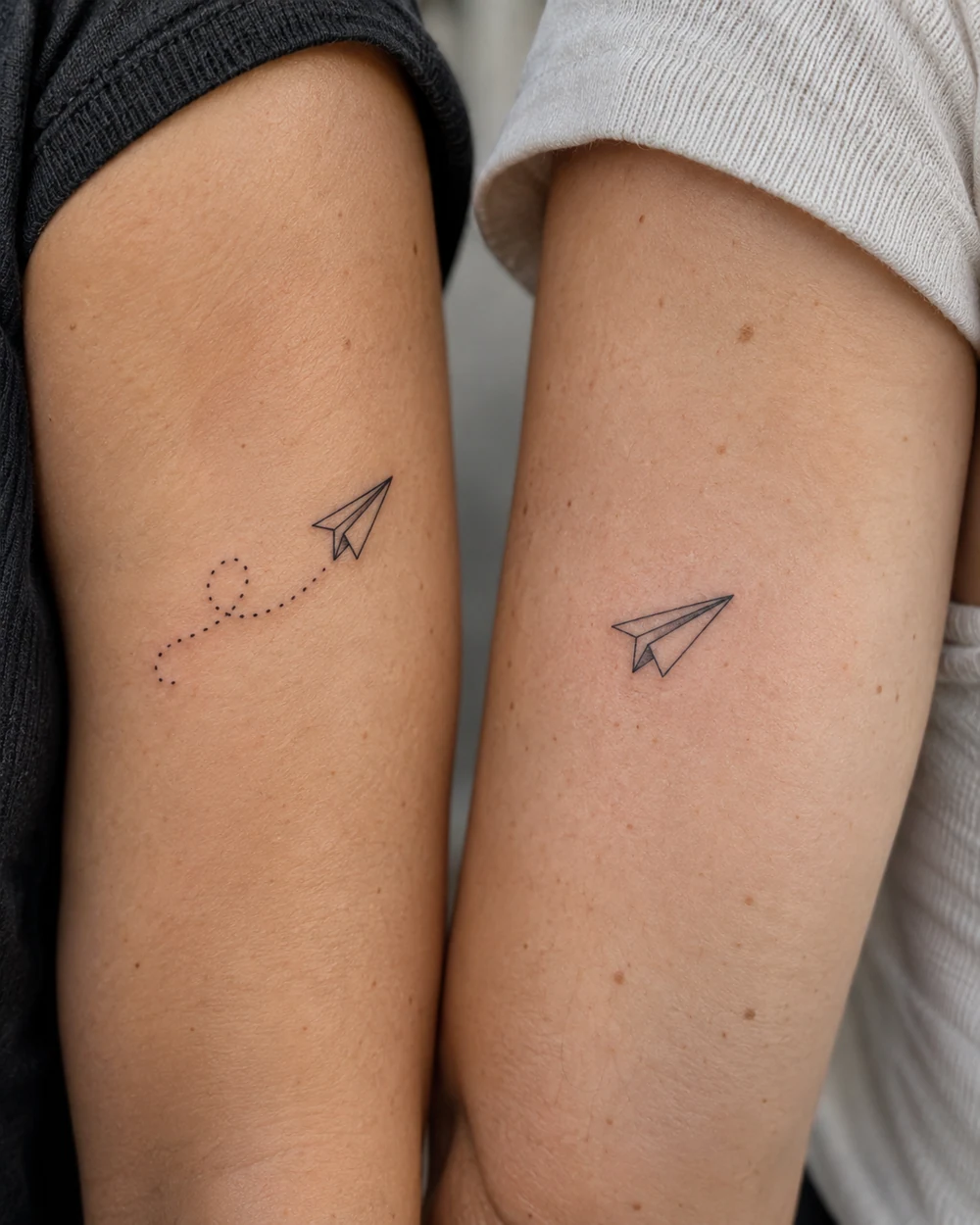 Matching fine-line paper-plane tattoos, one with a dotted trail, on two upper arms