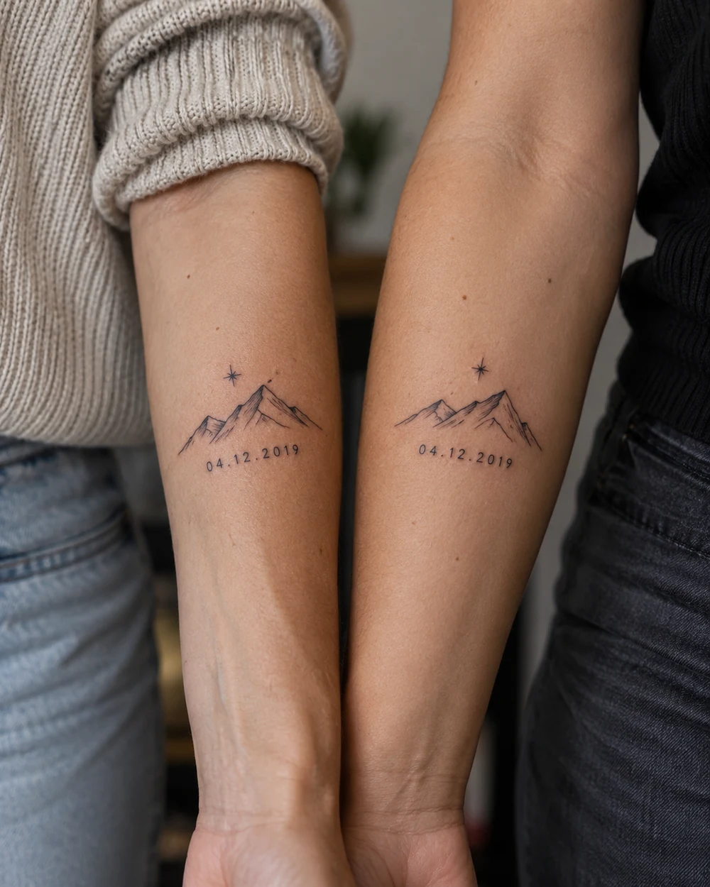 Two adult women’s forearms side by side, each bearing a matching fine-line mountain outline, star, and date tattoo.