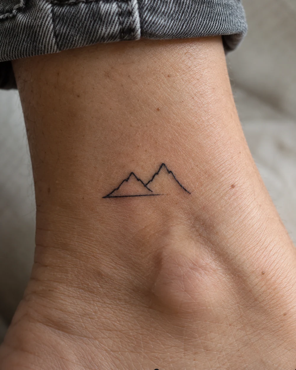 Fine-line black tattoo of two mountain peaks and a short baseline above the ankle
