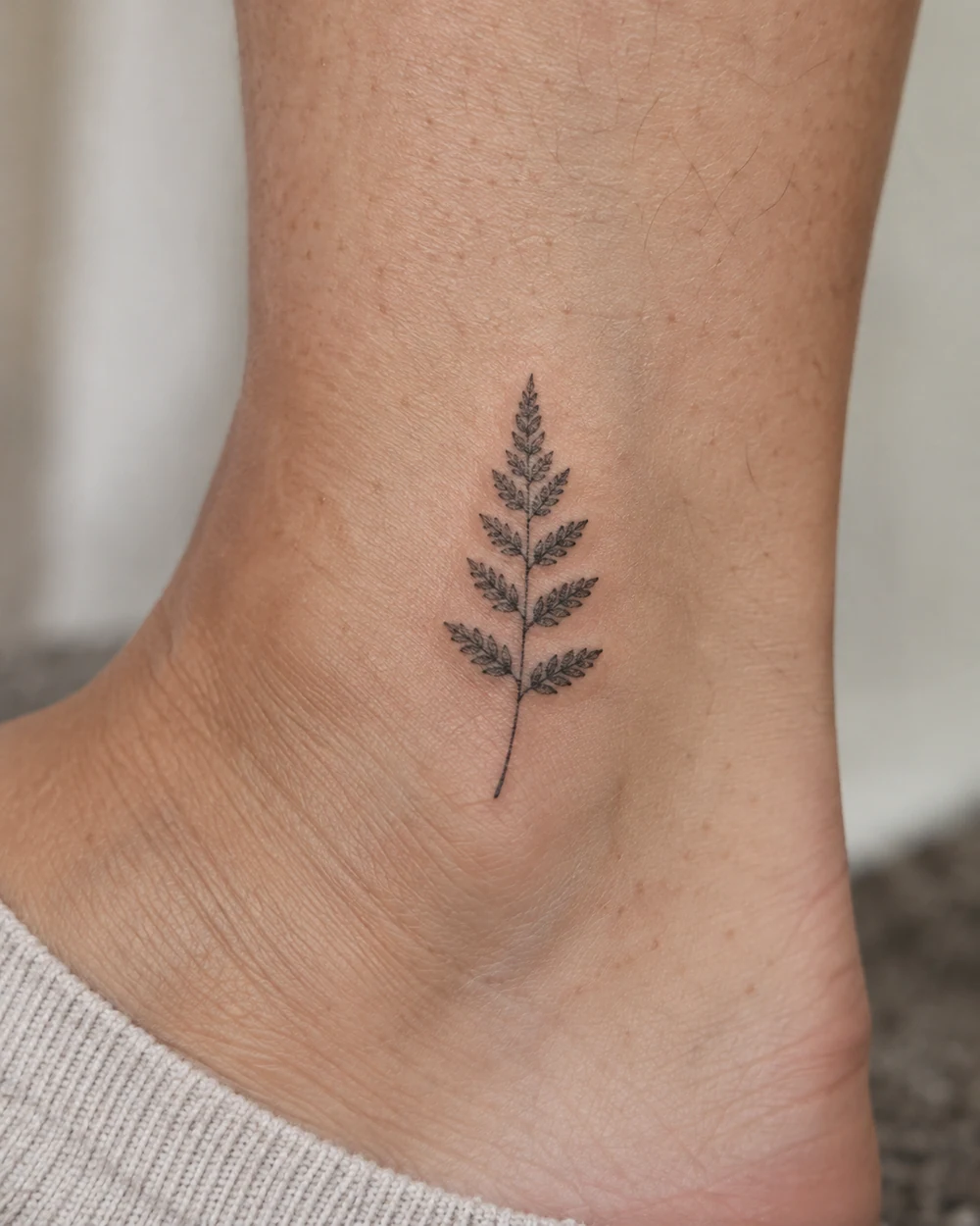 Fine-line black fern frond tattoo on an adult ankle