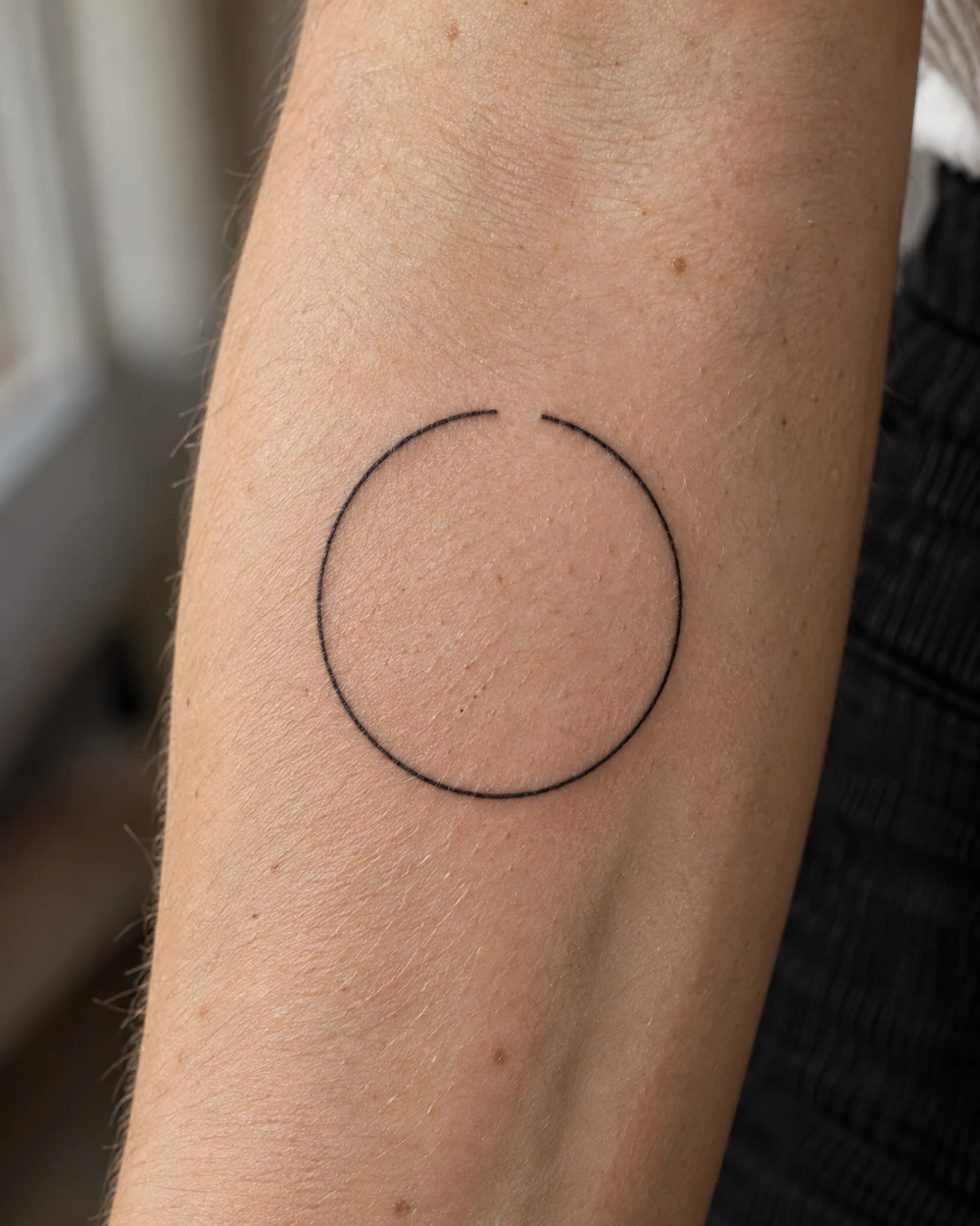 Fine-line black open circle tattoo on a forearm