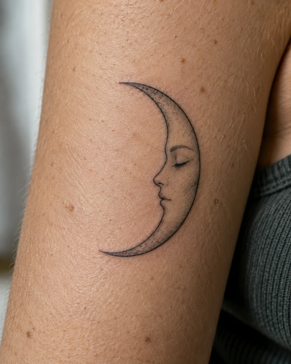 Fine-line black crescent moon tattoo containing a side-profile face with a closed eye