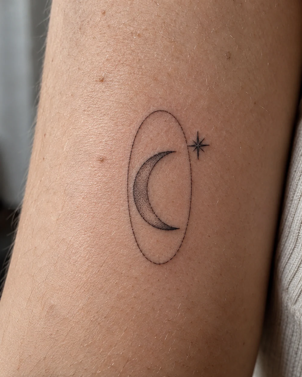 Fine-line crescent moon tattoo with stippled shading inside a narrow oval and a small star outside the oval