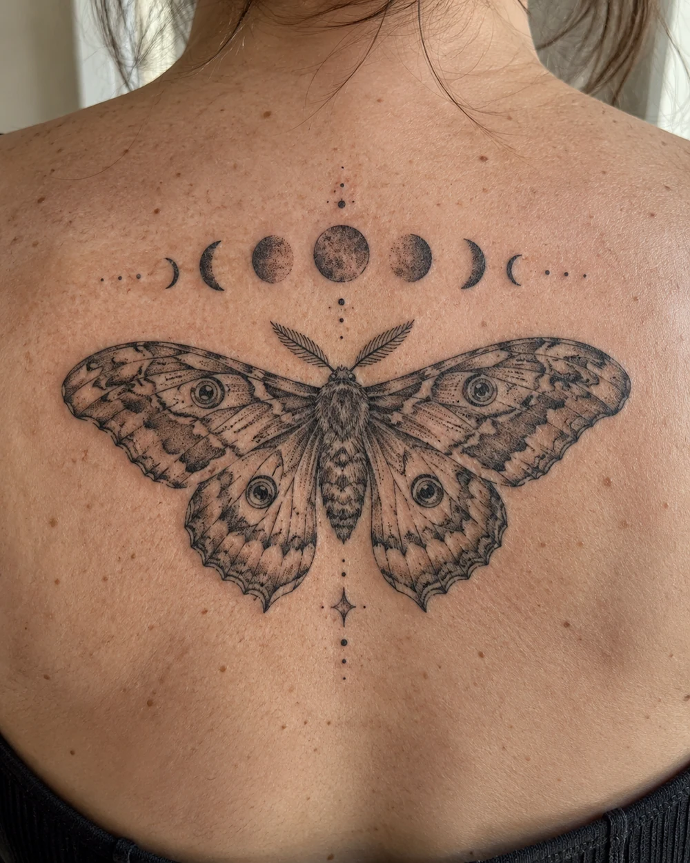 Black fine-line moth tattoo beneath moon phases and dots across an upper back