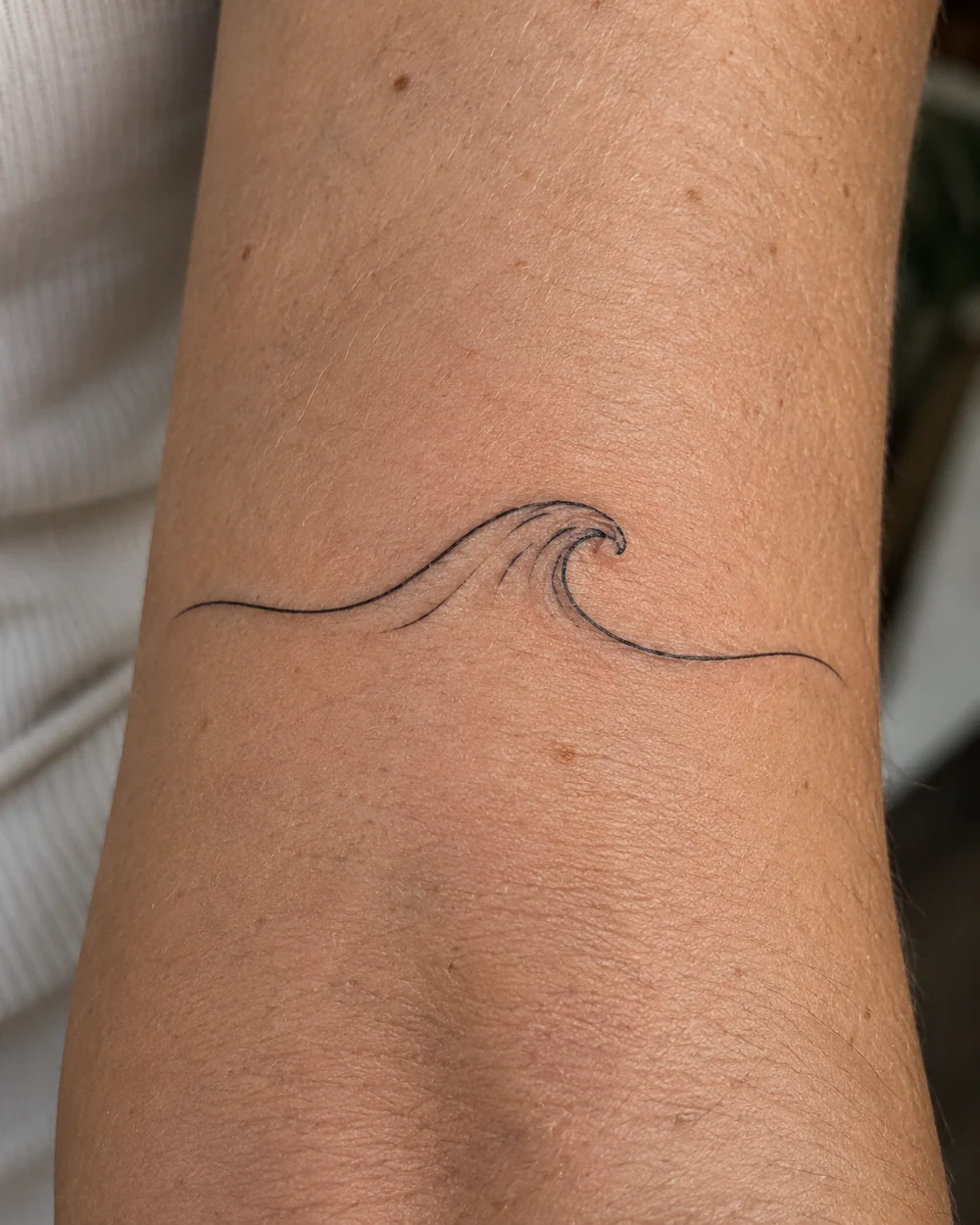 Minimal black continuous-line ocean swell tattoo with a small crest on the inner forearm