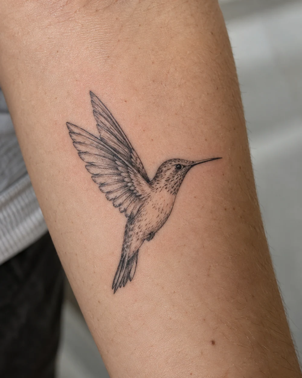 Fine-line hummingbird tattoo in flight on a forearm, with raised wings and narrow tail