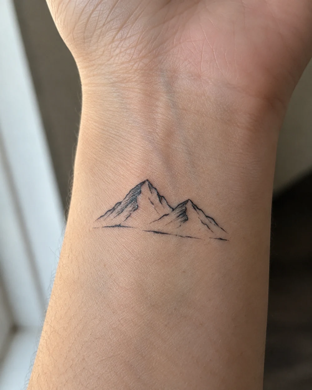 Small black mountain tattoo with two peaks, contour marks, and a short baseline on the wrist