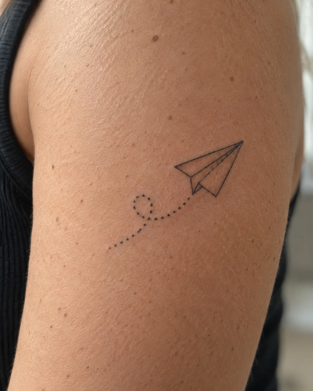 Fine-line paper plane tattoo with triangular folds and dotted trail on an upper arm
