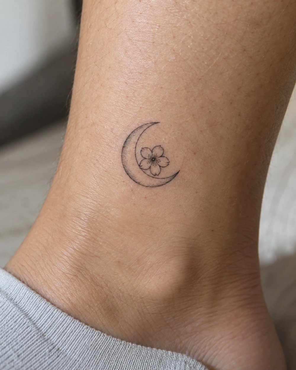 Small black fine-line crescent moon tattoo with a five-petal flower inside its curve, placed above the ankle