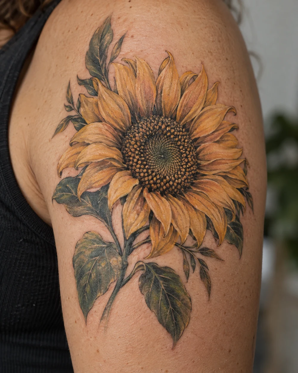 Golden-orange sunflower tattoo with a crisp seed-pattern center and dark green leaves on an upper arm