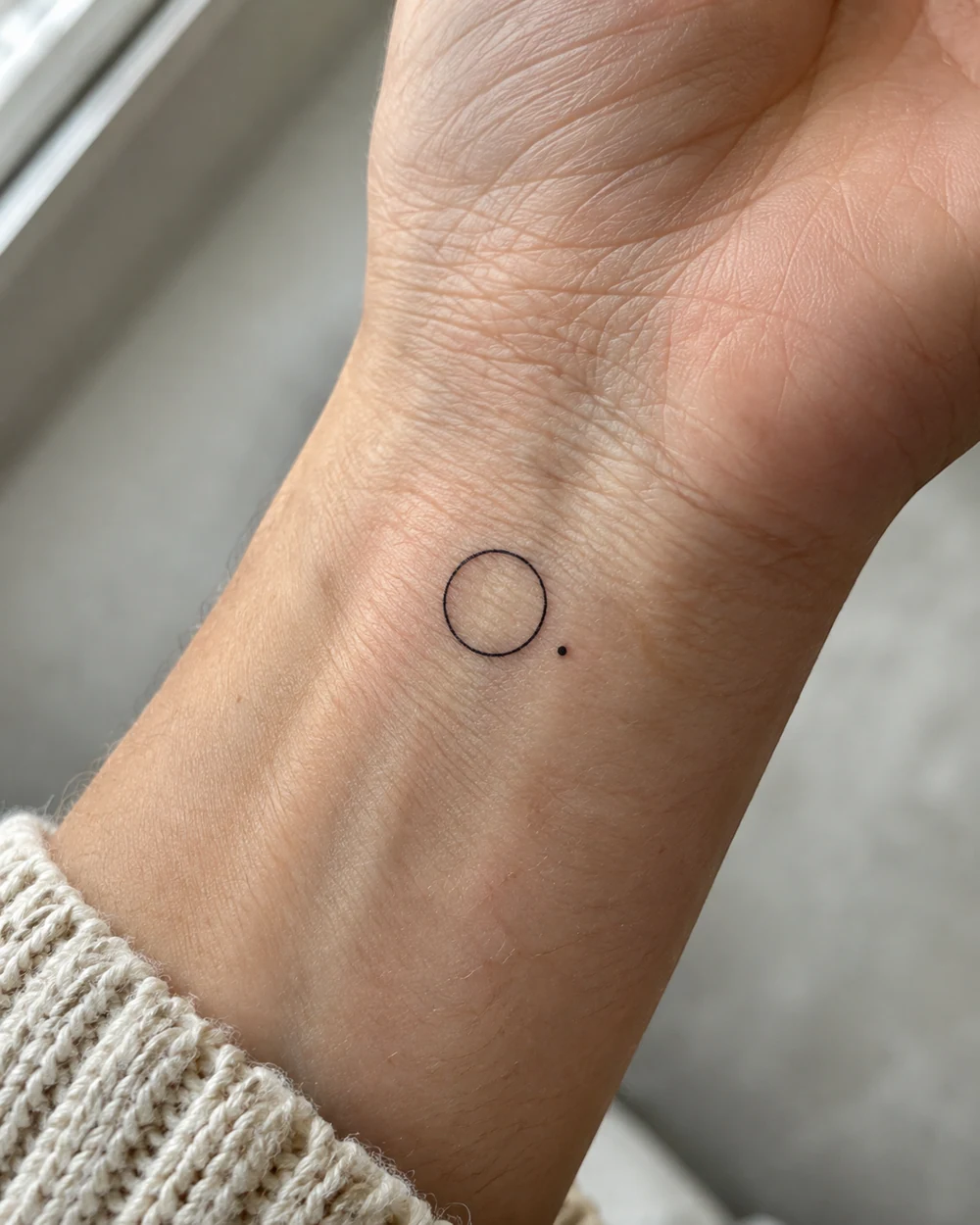 Small black open-circle tattoo with one dot on the inner wrist