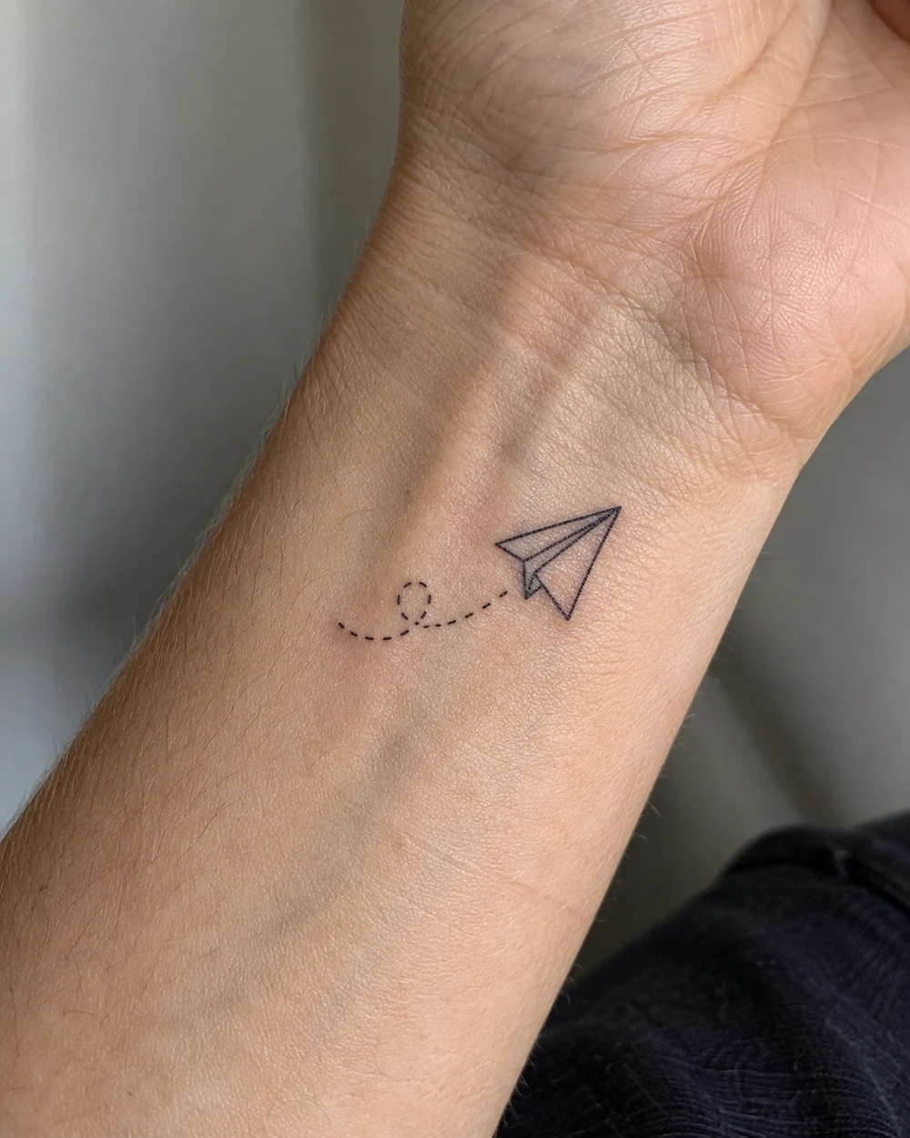 Tiny black paper-plane tattoo with a short dotted trail on a wrist